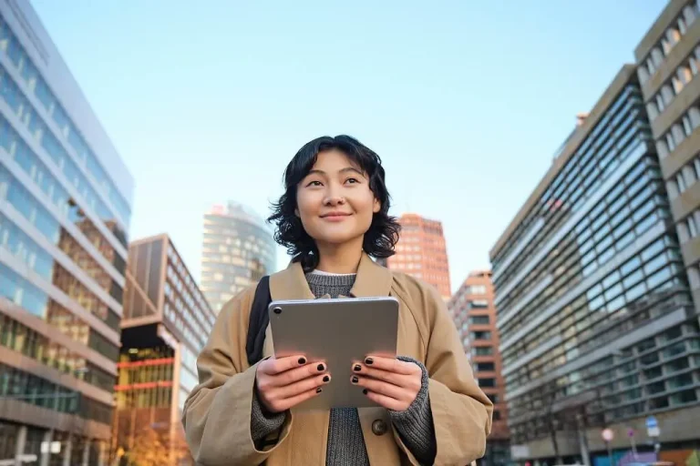 Portrait of a smiling Korean woman walking through the city with a digital tablet and backpack, enjoying an urban lifestyle.