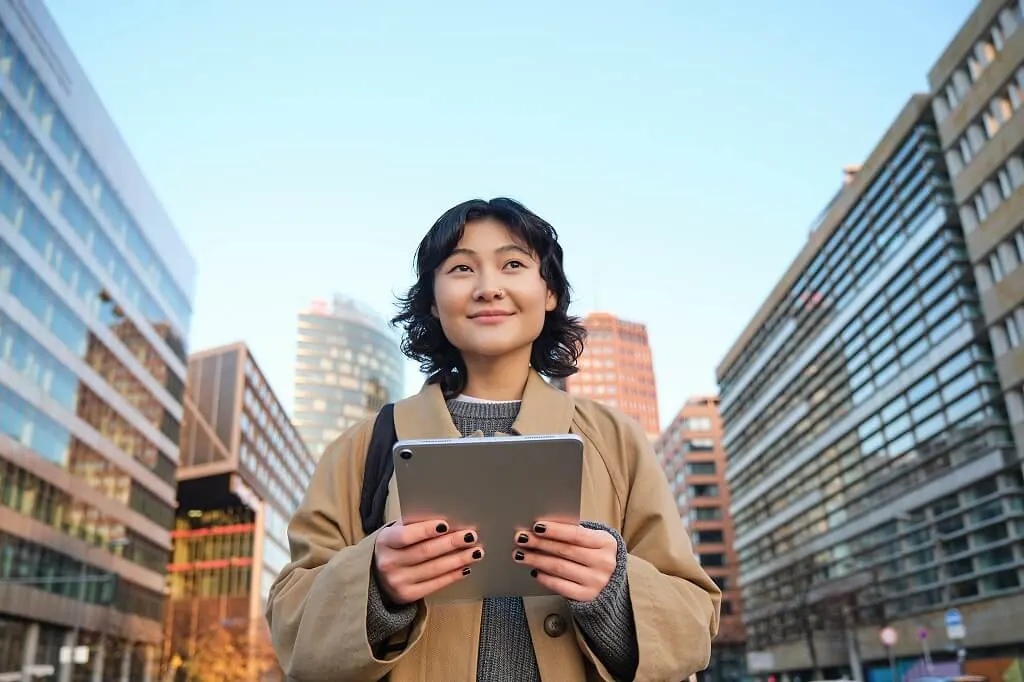 Portrait of a smiling Korean woman walking through the city with a digital tablet and backpack, enjoying an urban lifestyle.
